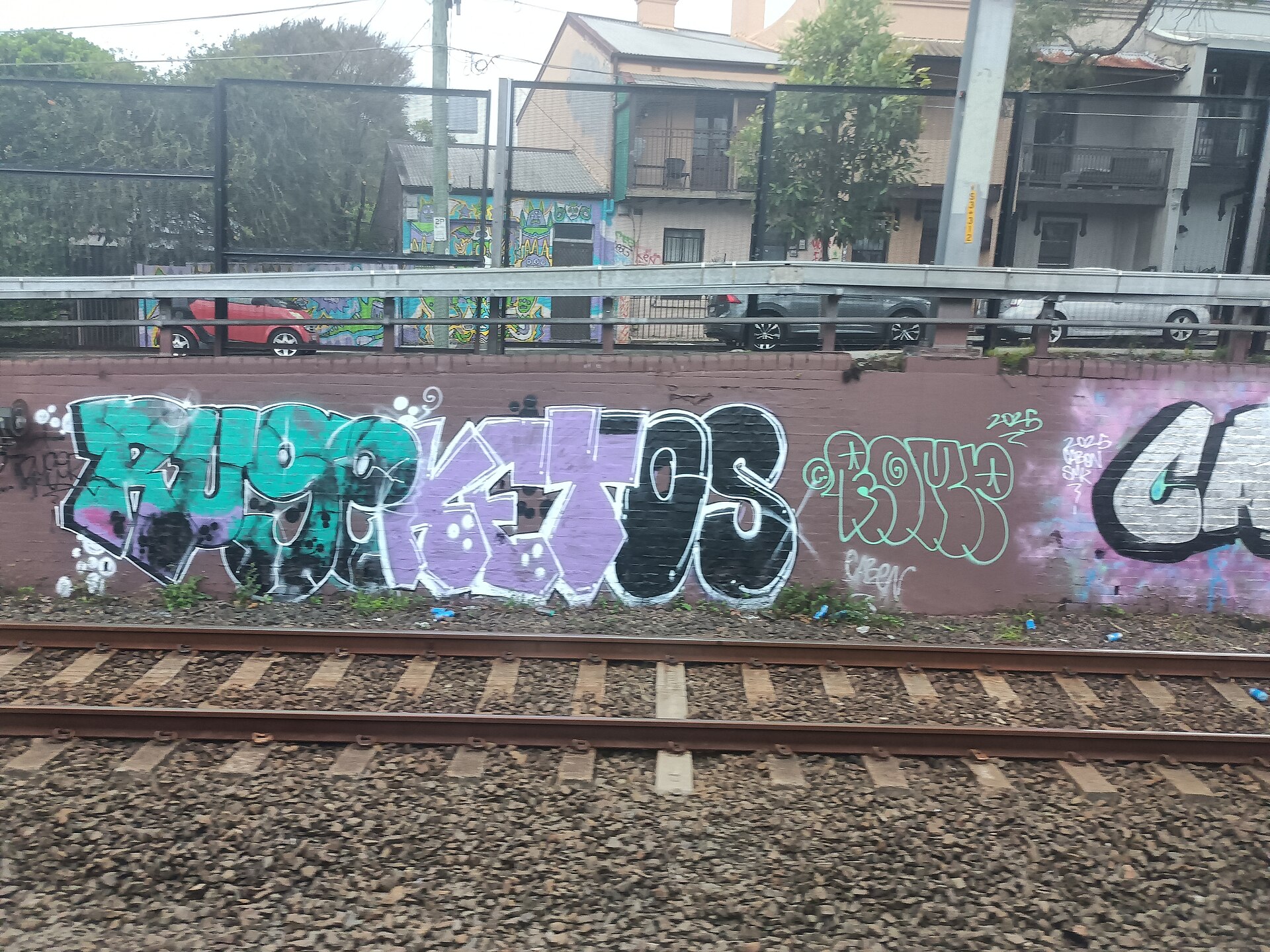 Teal, purple and black graffiti lettering along a brick wall beside a railway